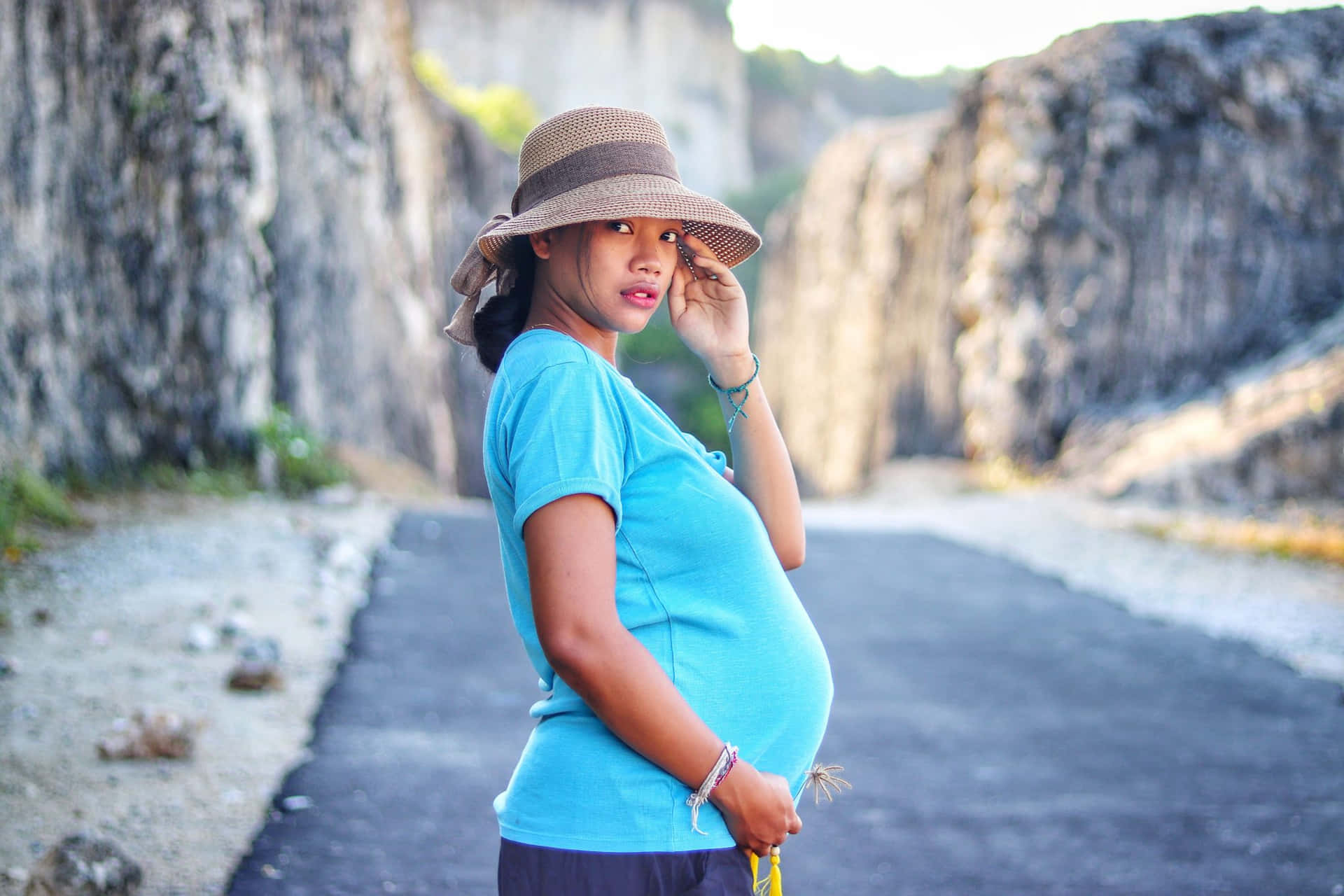 A Pregnant Woman In A Blue Shirt And Hat Standing On A Road