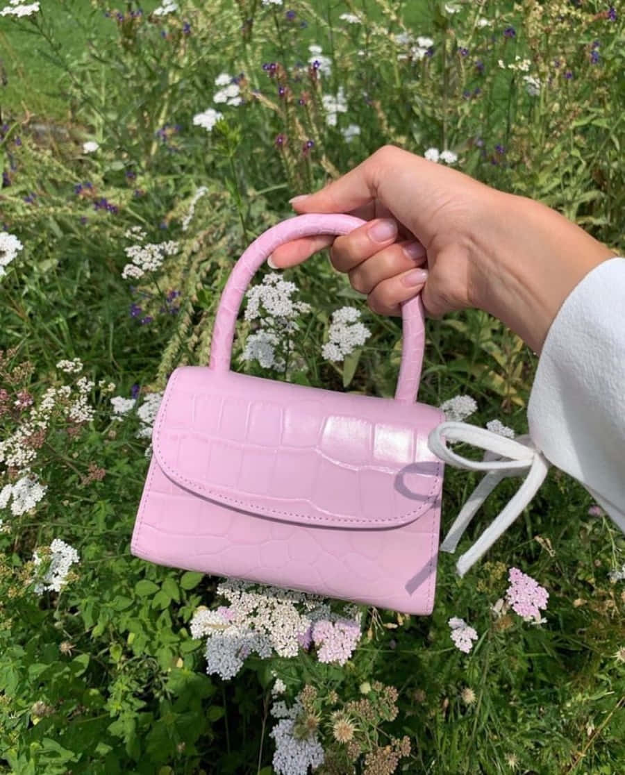 A Hand Holding A Pink Crocodile Purse In The Grass