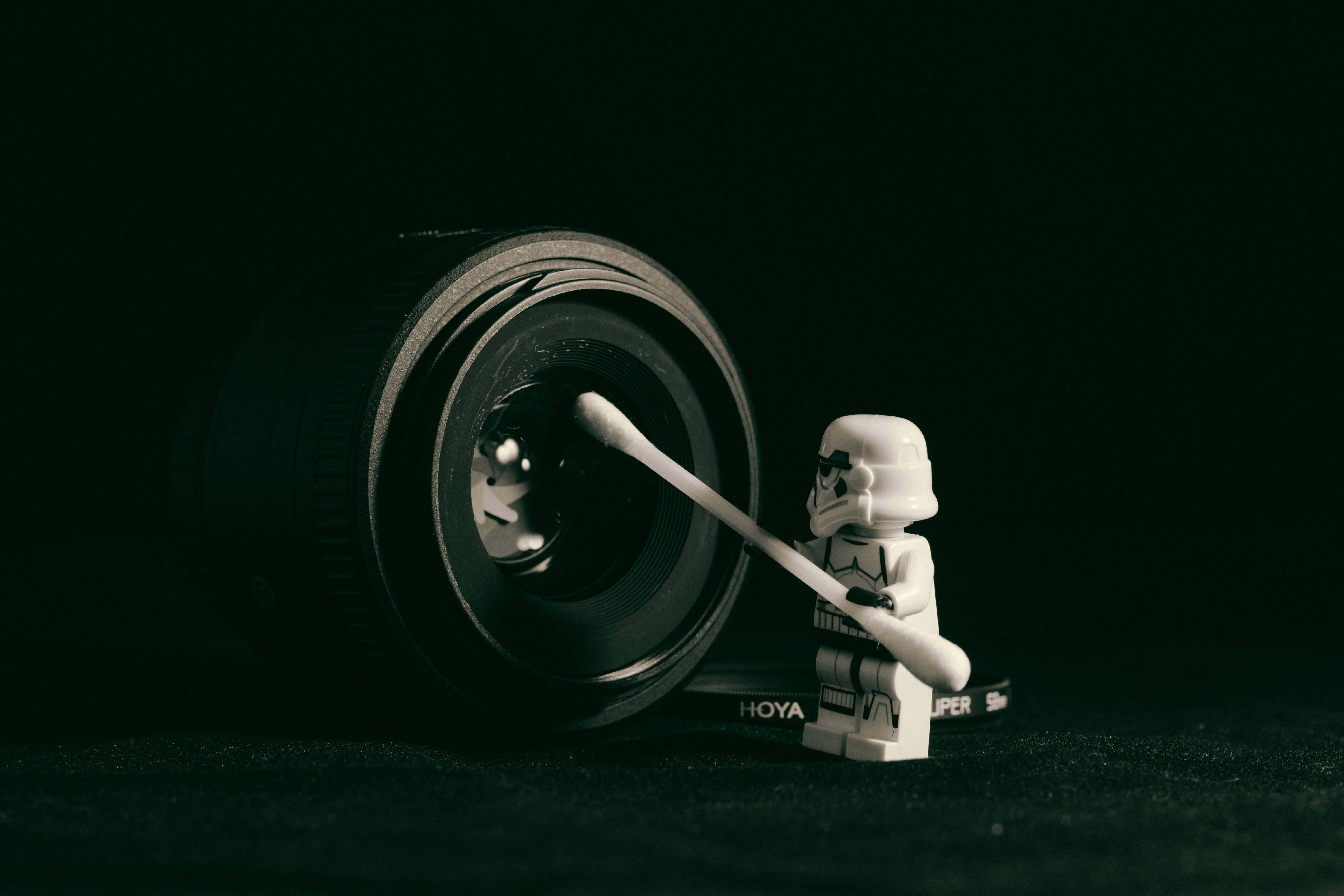 • “May The Laughs Be With You!” Wallpaper — A LEGO stormtrooper meticulously cleans a camera lens with a cotton swab against a stark black background.