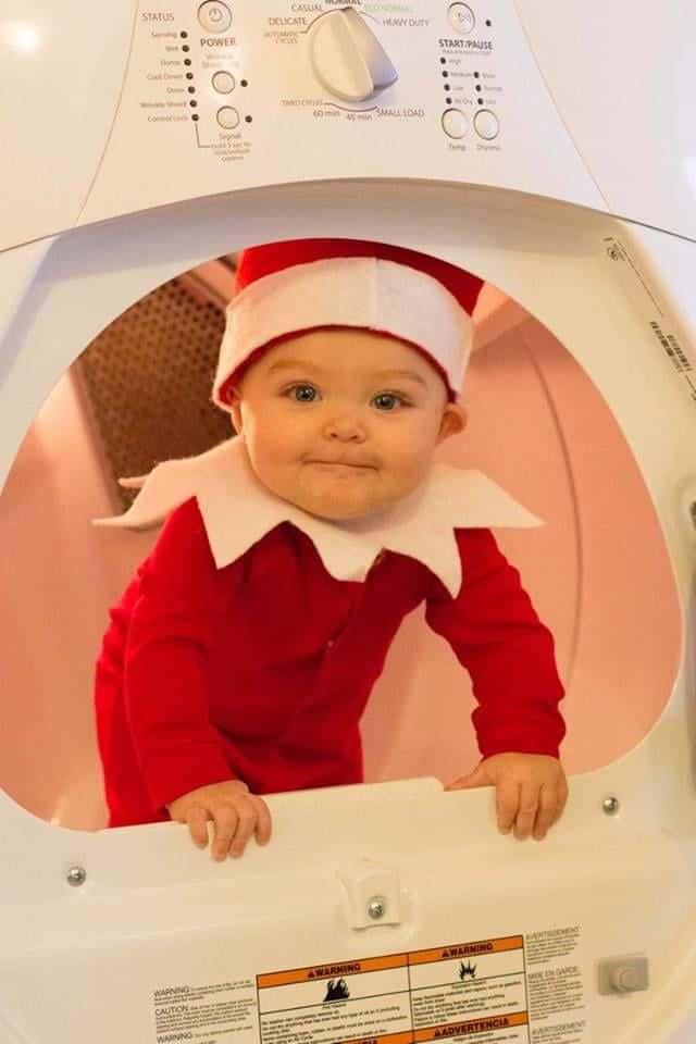 A Baby In An Elf Costume Is Looking Out Of A Washing Machine