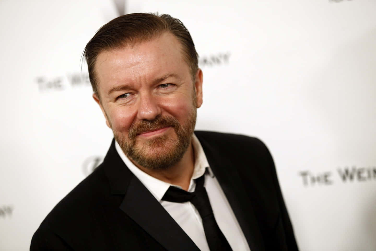Ricky Gervais, adorned with a smart casual look, smiling at the camera. Wallpaper