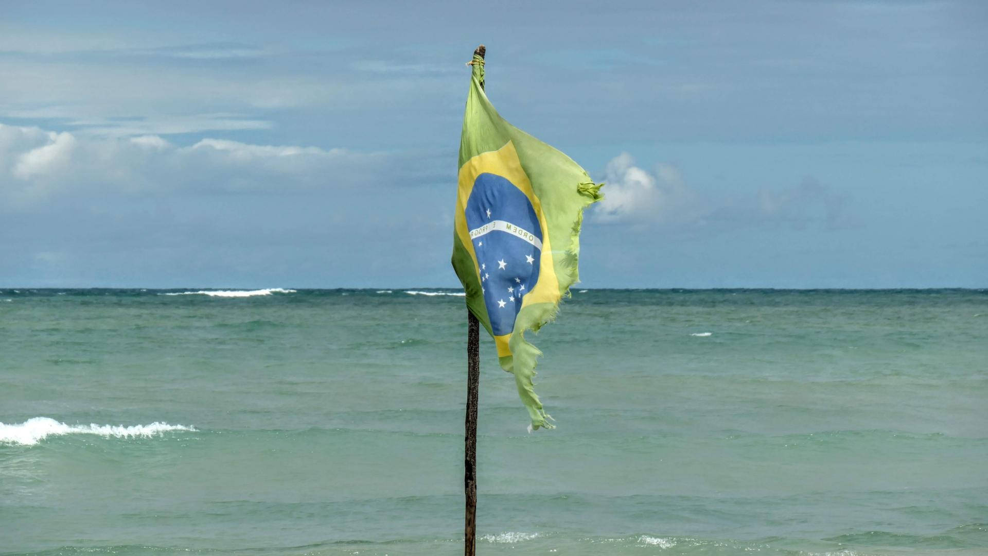A Majestic Brazilian Flag Fluttering Against a Beautiful Beach Landscape Wallpaper
