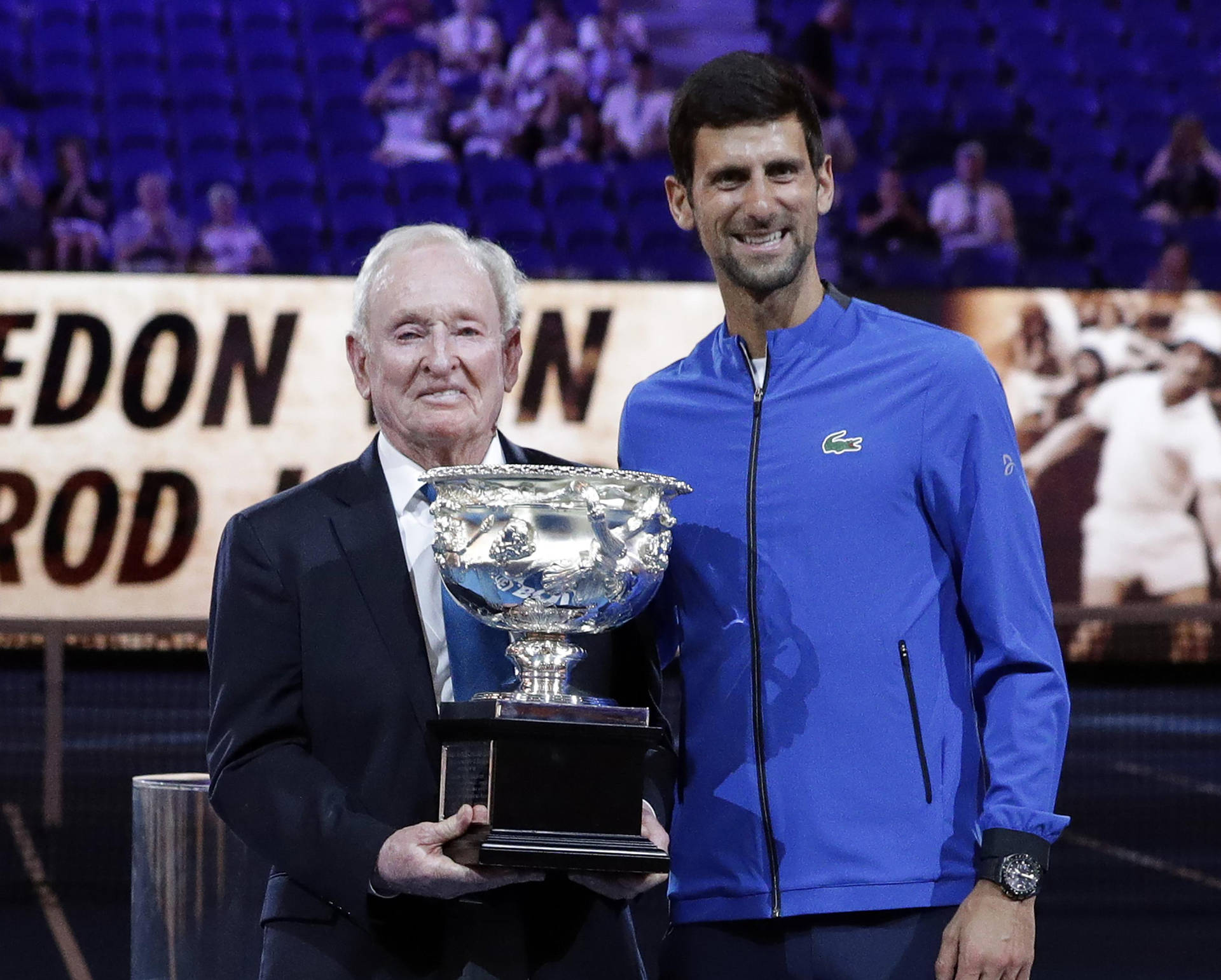 Rod Laver Holding Trophy With Novak Djokovic Background