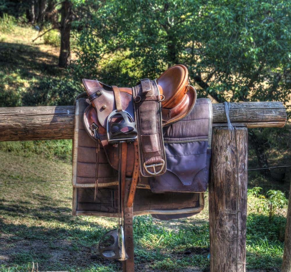A brown leather saddle resting on a wooden fence against a rustic countryside background Wallpaper