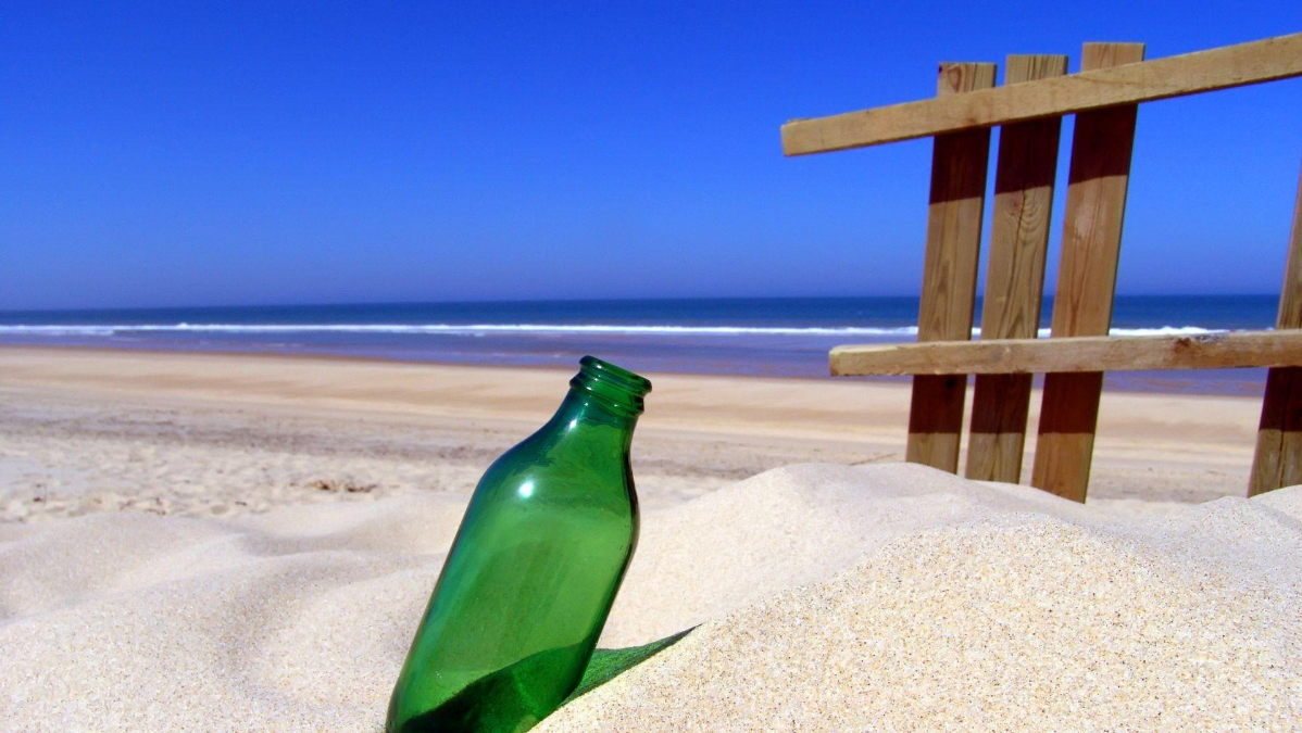 A layer of golden sand drenched with the setting sun's last rays Background — A green glass bottle rests in sand beside a wooden fence on a bright sunny beach with ocean waves and clear blue sky.