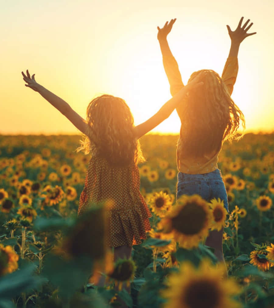 Two Girls Standing In A Sunflower Field At Sunset