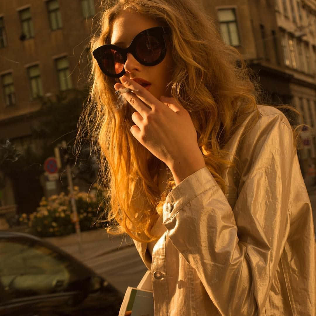 A Woman Wearing Sunglasses Smoking A Cigarette In The Street