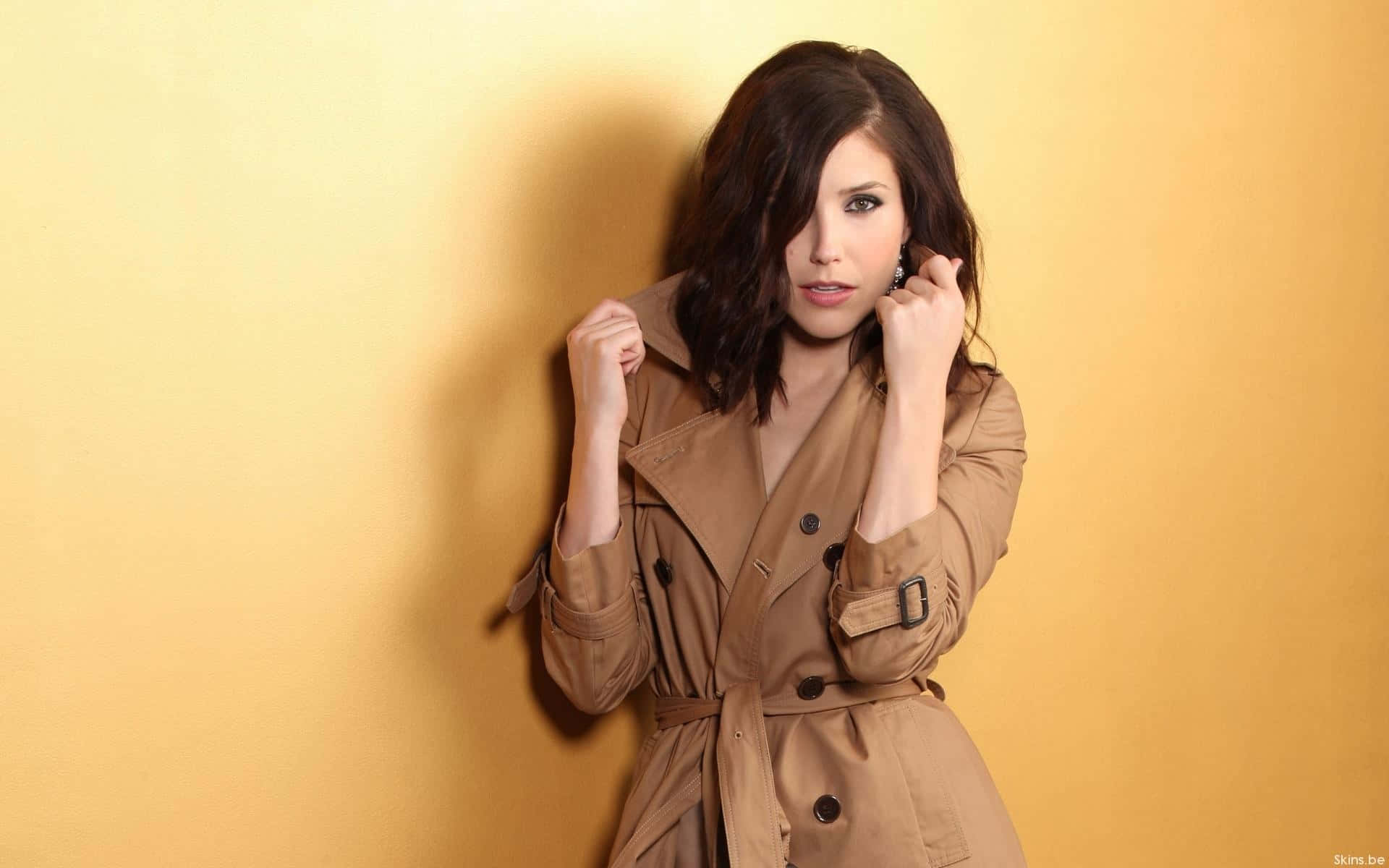 A Woman In A Trench Coat Leaning Against A Yellow Wall Wallpaper