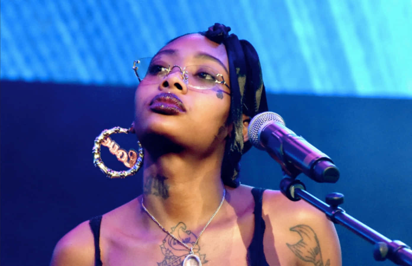 A Woman With Tattoos And Glasses Singing Into A Microphone