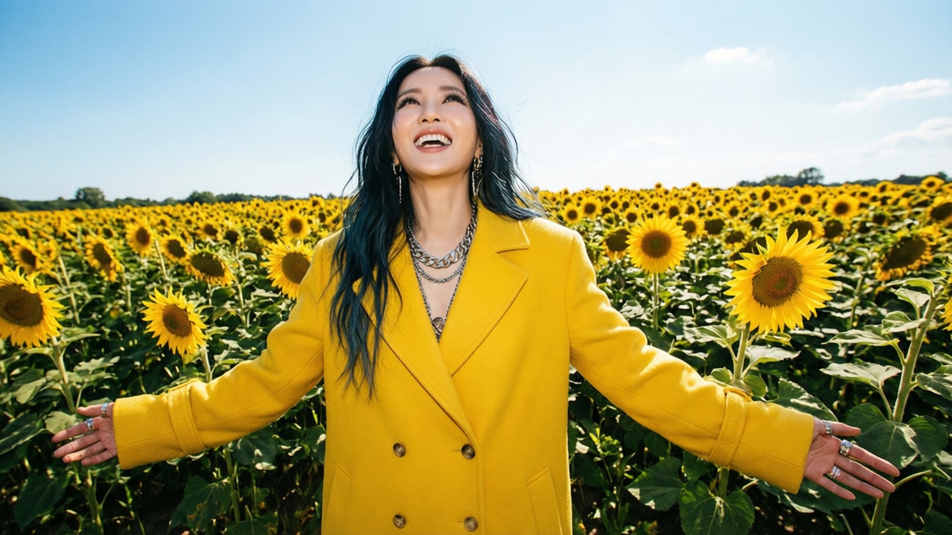 Champs de tournesols Fonds d'écran — Hayley Kiyoko dans un manteau jaune parmi les tournesols, bras ouverts.