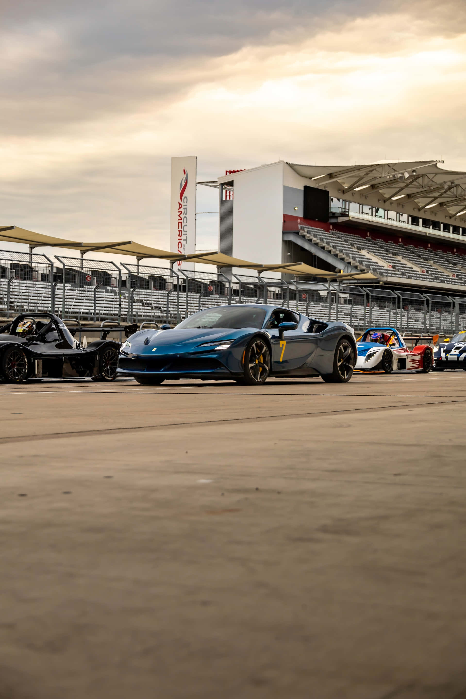 Supercars Wallpaper — A sleek blue race car with number 7 parked on a racetrack beside other sports cars under a cloudy sky.