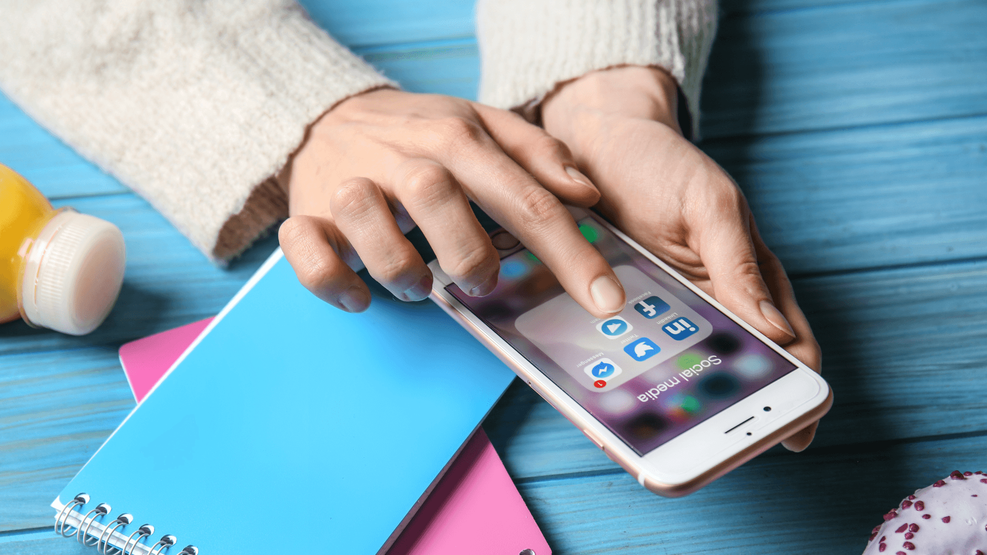 Stay connected with friends, colleagues and other contacts - message, call, and share with Telegram! Background — Hands scroll social media apps on a rose gold phone resting on colorful notebooks and a blue wooden table.