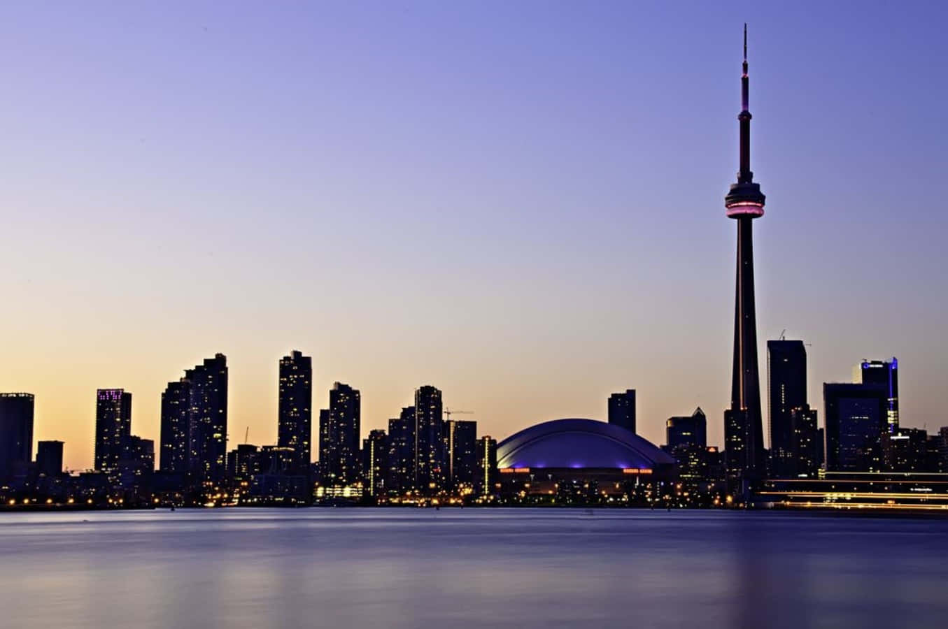 The glowing Toronto skyline reflects in the calm evening waters.