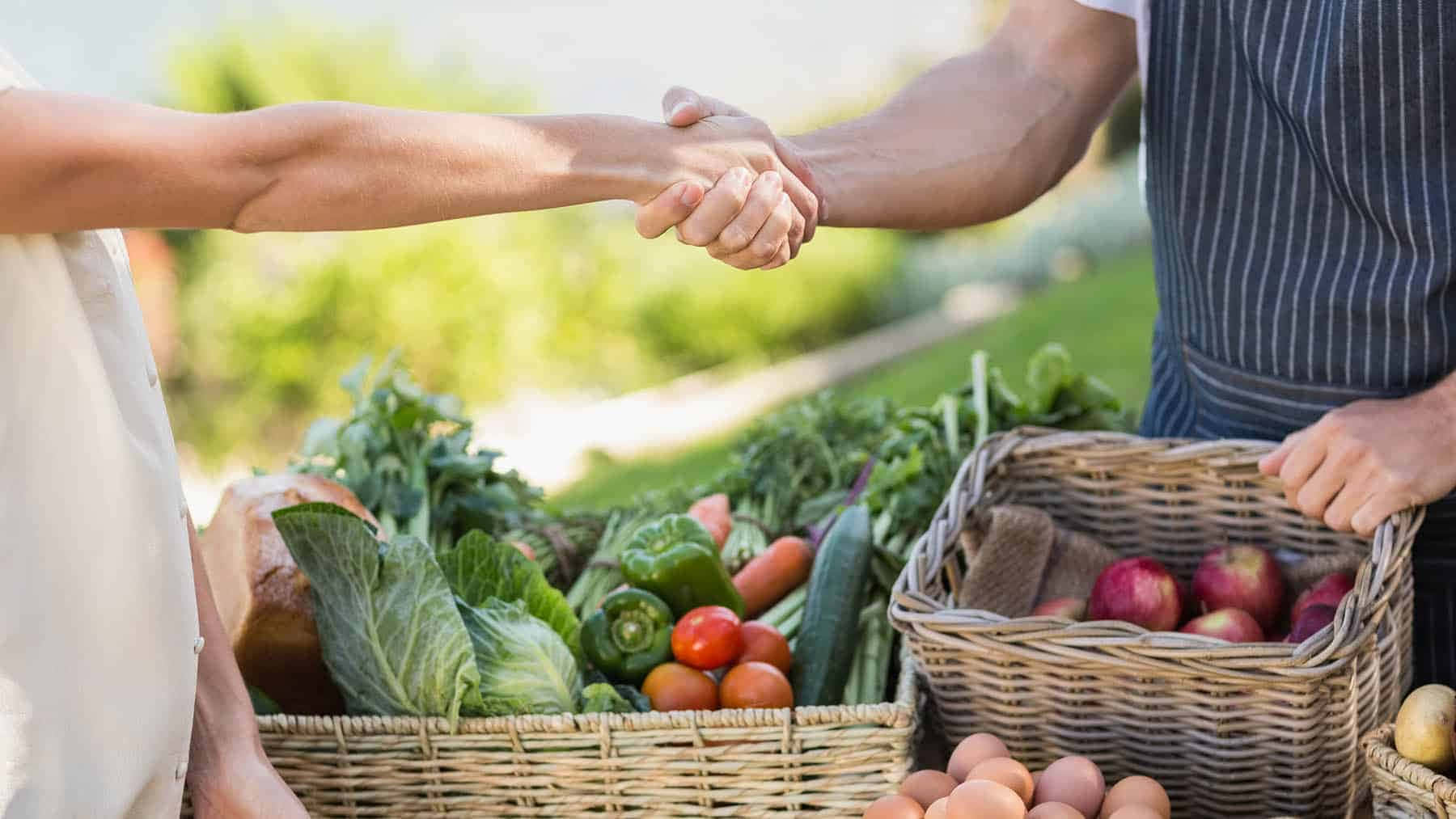 A Man And Woman Shaking Hands In Front Of A Basket Of Vegetables
