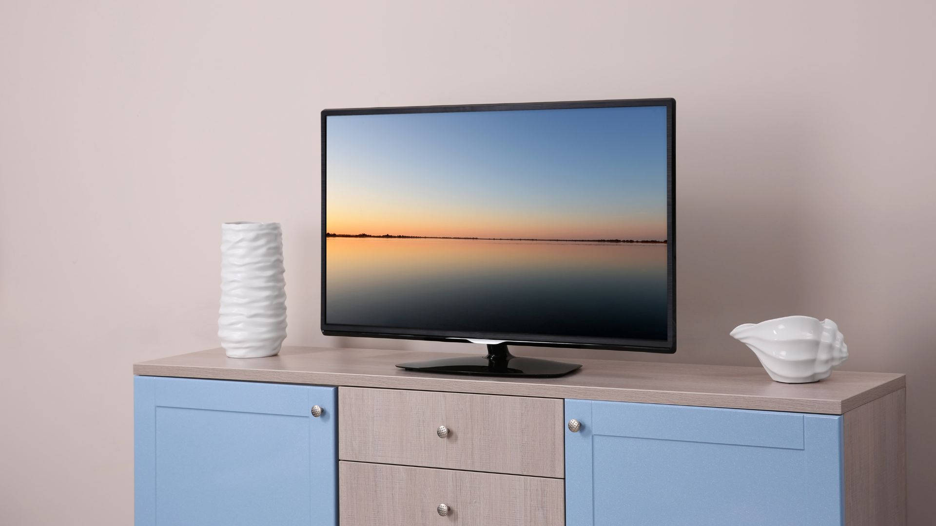 32 Inch LED Full HD TV Wallpaper — A modern TV displaying a serene sunset over water sits atop a light wood cabinet with blue doors and white ceramic decor.