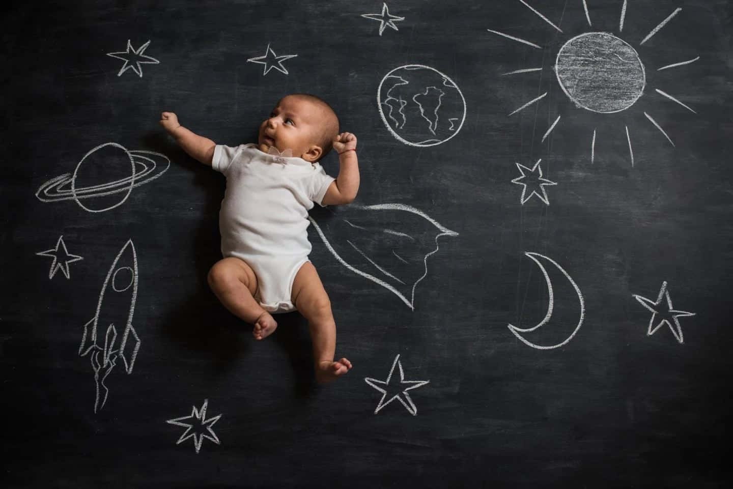 A Baby Laying On A Chalkboard With Stars And Planets Drawn On It