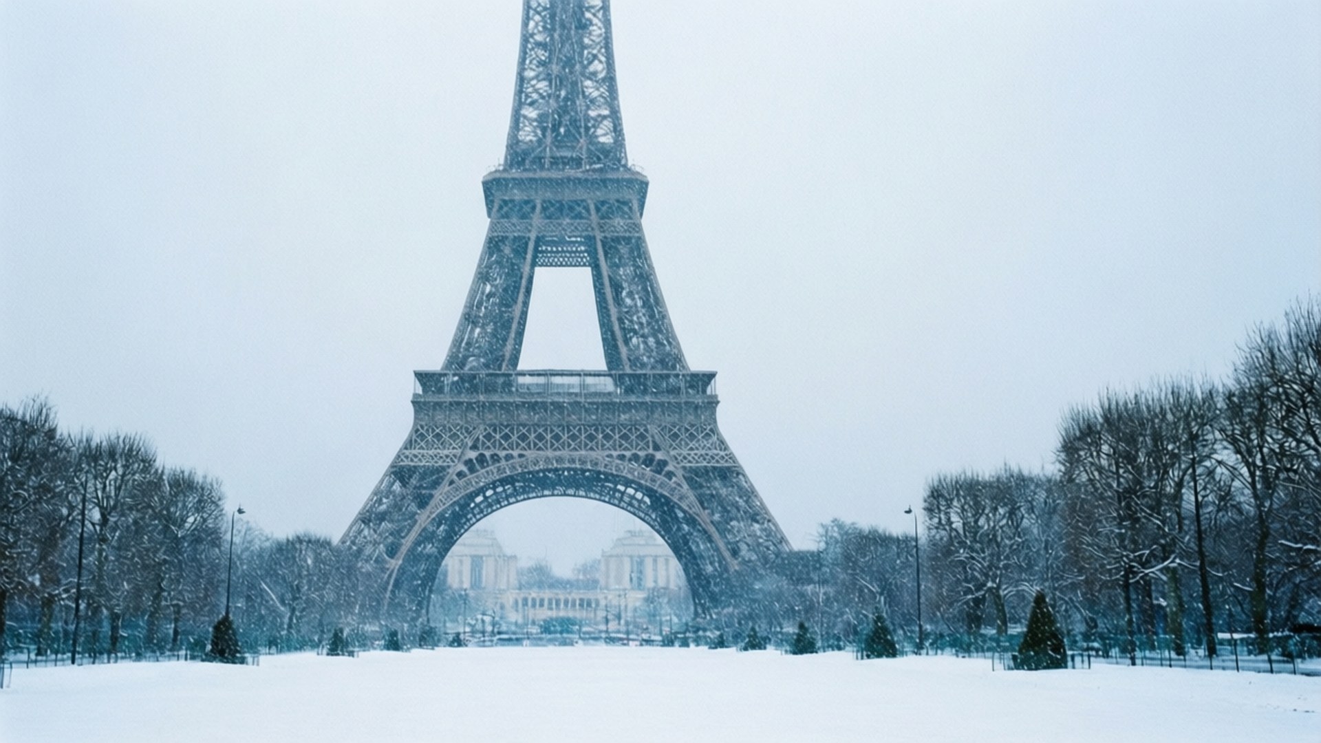 Winter Wonderland Eiffel Wallpaper — The Eiffel Tower stands majestic under a snowy Parisian sky with bare trees and a blanket of white snow covering the ground.