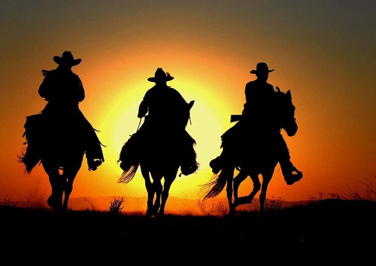 Silhuet af tre cowboys der rider på heste mod en dramatisk solnedgang Wallpaper
