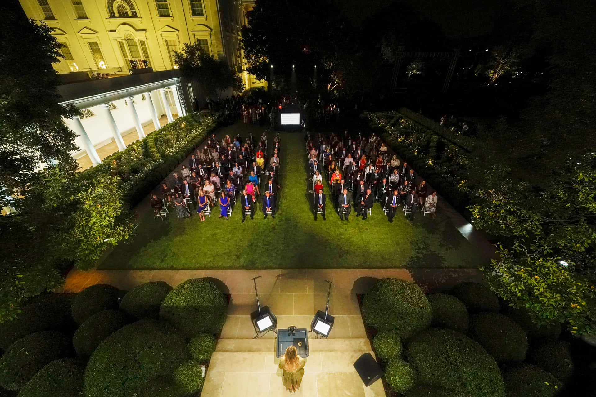 A Crowd Of People Standing In A Garden At Night