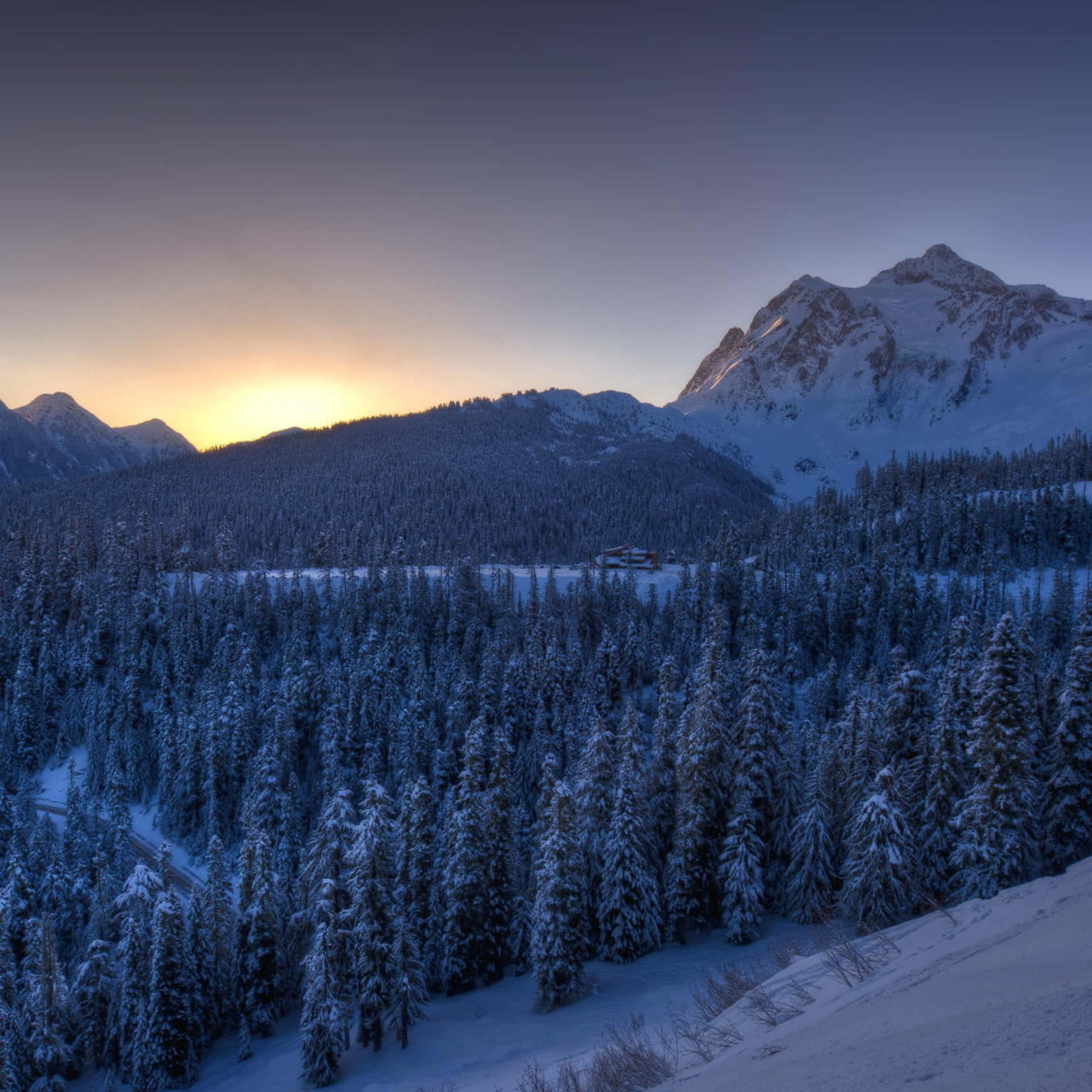 A Snowy Mountain Range With Trees And A Sun Setting Wallpaper