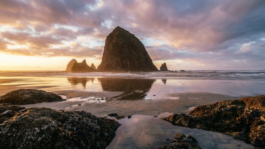 Ikoninen Haystack Rock Cannon Beachillä auringonlaskussa vuorovesialtaiden ja dramaattisten pilvien kanssa.