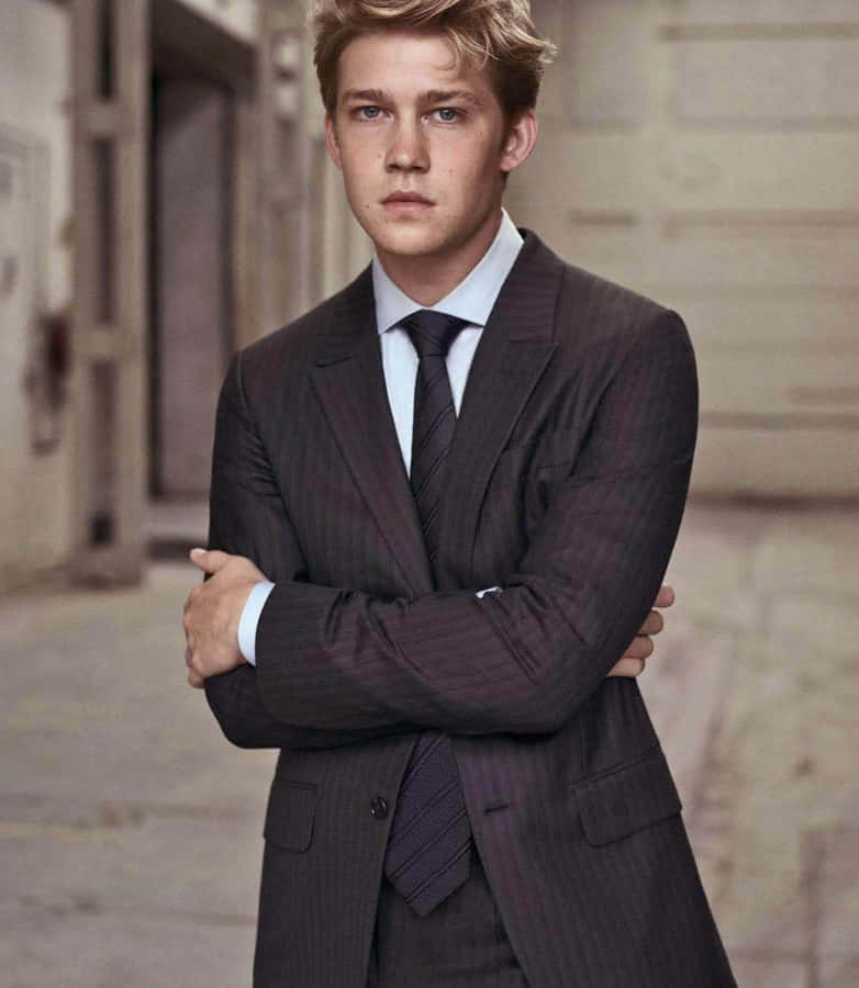 Elactor inglés Joe Alwyn vistiendo un traje gris con un fondo azul brillante.