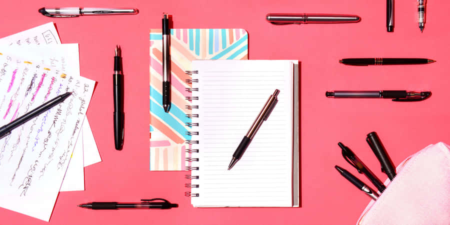 EinNotizbuch, Stift, Bleistift und andere Schreibutensilien auf einem rosafarbenen Hintergrund.