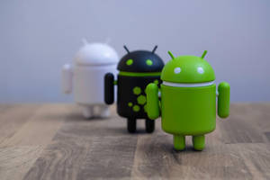 Três bonecos robôs Android nas cores verde, preto e branco sobre uma superfície de madeira. — Desenvolvedor Android Papéis de Parede