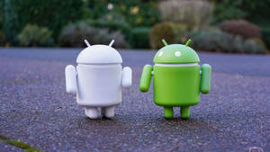 Duas figuras de robô Android, branca e verde, em pé em caminho de cascalho ao ar livre — Desenvolvedor Android Papéis de Parede