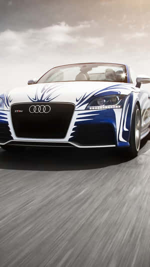 Audi TT RS decappottabile bianca e blu che sfreccia su una strada grigia sotto cielo nuvoloso.