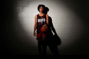 Junger Basketballspieler in Huskies-Trikot hält Ball unter Scheinwerferlicht vor Ziegelwand. — Jared Mccain Wallpaper