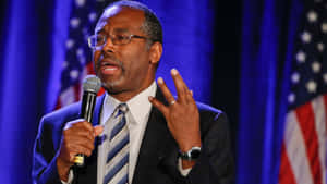 Man in suit speaking into microphone with American flags behind him.