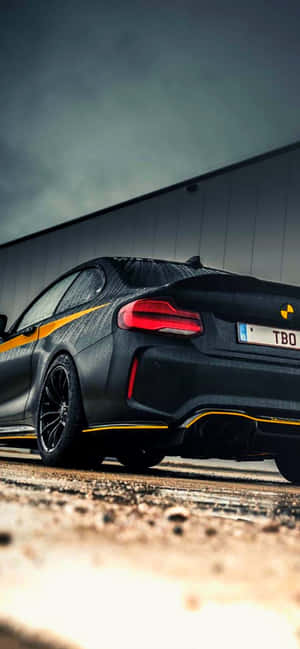 Black sports car with yellow stripes parked on gravel near industrial building. — Bmw Android Picture