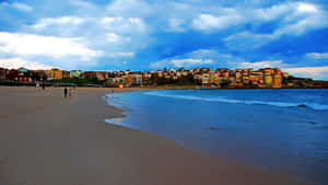 Two people stroll along a sandy beach at dusk with colorful houses on the hillside.