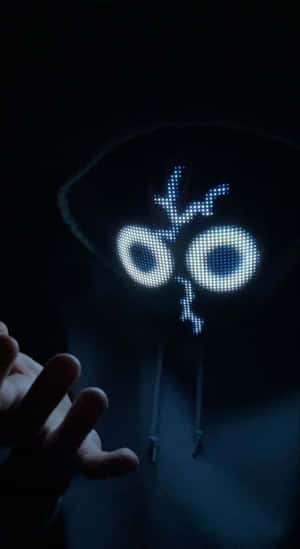 Eine Person in einem dunklen Kapuzenpullover trägt eine leuchtend blaue LED-Maske in Form eines Cartoon-Gesichts.