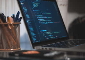 Laptop with code on screen next to a pen holder on a desk. — Code Wallpaper