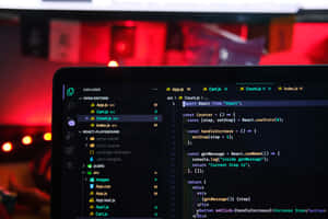 Laptop screen showing React code with red ambient lighting in a tech workspace. — Code Wallpaper