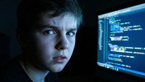 Ung dreng intenst fokuseret på kodning på en lysende computerskærm i et mørkt rum.