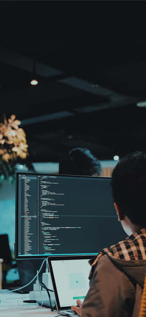 Pessoa programando em laptop e monitor em escritório com pouca luz à noite. — iPhone com padrão fofo Papéis de Parede