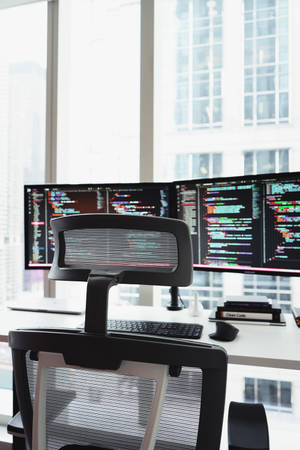 Coderens arbejdsstation med tre skærme med kode og byudsigt gennem vinduet.