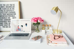 Uma mesa branca arrumada com laptop, rosas cor-de-rosa, candeeiro dourado e arte com citação motivacional. — Computador portátil Sunflower Papéis de Parede