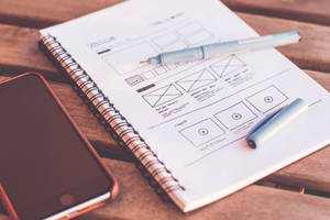 Anteckningsbok med wireframe-skisser och pennor på ett träbord bredvid en smartphone. — Cool iPhone Bakgrundsbilder