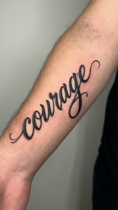 Een elegante schrift-tattoo met de tekst 'moed' op een onderarm.