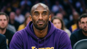 Kobe Bryant duduk di pinggir lapangan dengan hoodie ungu, tenang dan reflektif, dengan latar belakang arena yang kabur lembut.