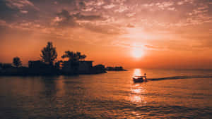 A boat sails on water during a vibrant orange sunset with silhouetted trees and buildings.
