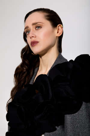Woman in black ruffled jacket and gray herringbone top with wavy hair and bold makeup.