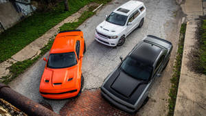 Três Dodge Challengers estacionados em uma rua rachada com bordas gramadas. — Esquivar Papéis de Parede