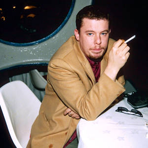Man in tan coat holding cigarette at table in dim interior — Funny Mario Picture
