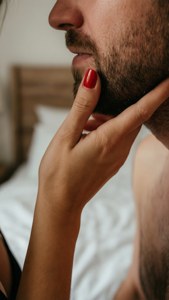 De hand van een vrouw houdt zachtjes het gezicht van een man vast, rode nagels, warm licht, bezitterig gebaar.