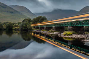 A speeding train crosses a green bridge over a river with mountain reflections. — Google Landscape Background